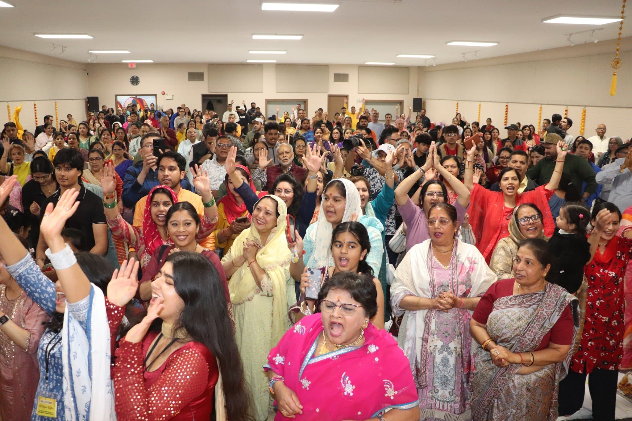 A packed hall of devotees singing with hands raised in devotion